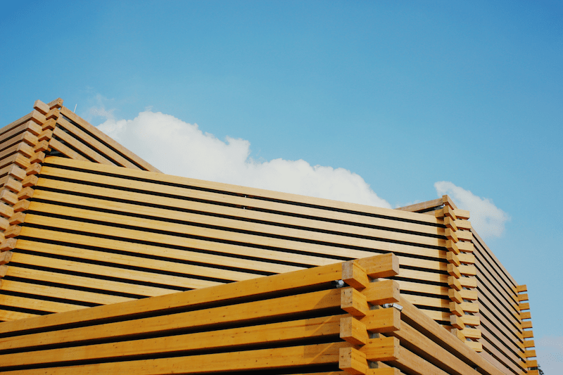Moderne Holzkonstruktion aus hellen Holzbalken in rechteckiger FormBauweise von unten gesehen, im oberen Bereich blauer Himmel mit einzelnen Wolken