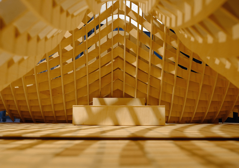 Eine kunstvolle geschwungene Holzkonstruktion im Hintergrund, davor heller Holzboden und ein stützendes Holzelement