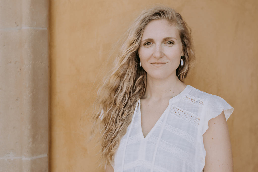 Porträt-Foto einer lächelnden Frau mit blondem, langen Haar vor einer erdfarben gestrichenen Wand